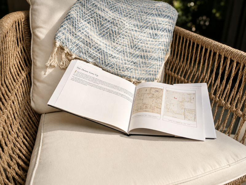 A Deed & Dwelling house history book lying open to a chapter titled 'The House Goes Up,' resting on an outdoor chair